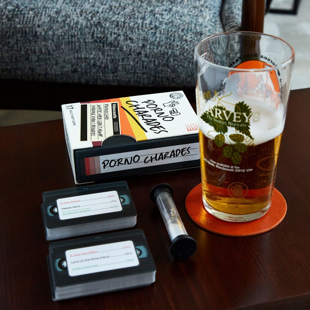 Glass of beer on a coaster with &#39;Harvey&#39; branding, a box of &#39;Porno Charades&#39;, and game cards on a table.