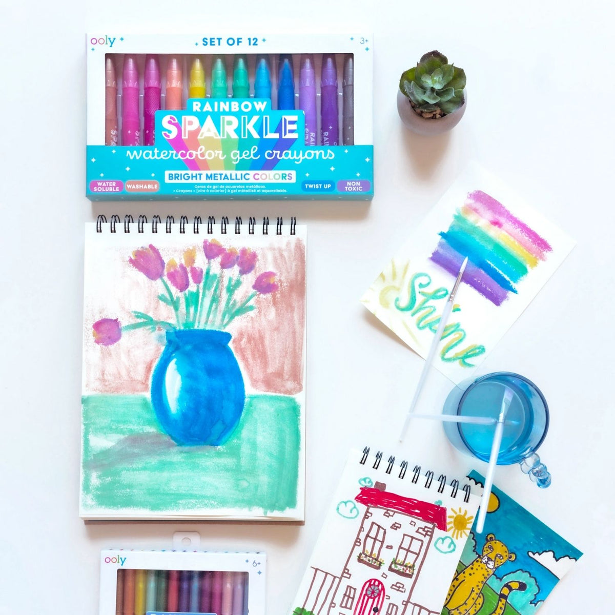 A set of 12 rainbow sparkle watercolour gel crayons by ooly, with each crayon featuring a metallic color. The packaging displays the crayons in a row with a label indicating the product is washable and has a twist-up mechanism.