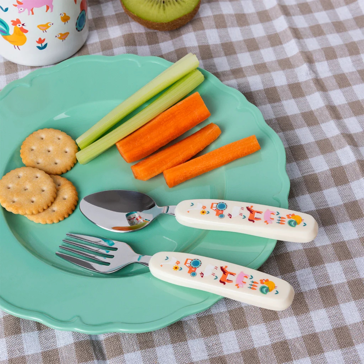 Children&#39;s plate with crackers, carrots, celery, and floral-patterned fork and spoon on a checkered tablecloth.
