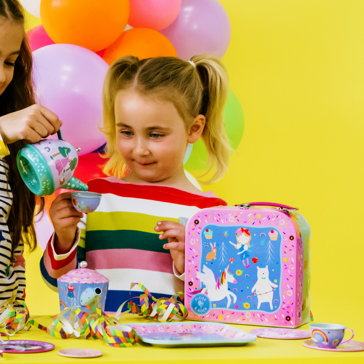 two girls playing with A Rainbow Fairy themed tin tea set in a colorful box with illustrations of fairies and animals. The set includes a teapot, cups, saucers, and a tray.