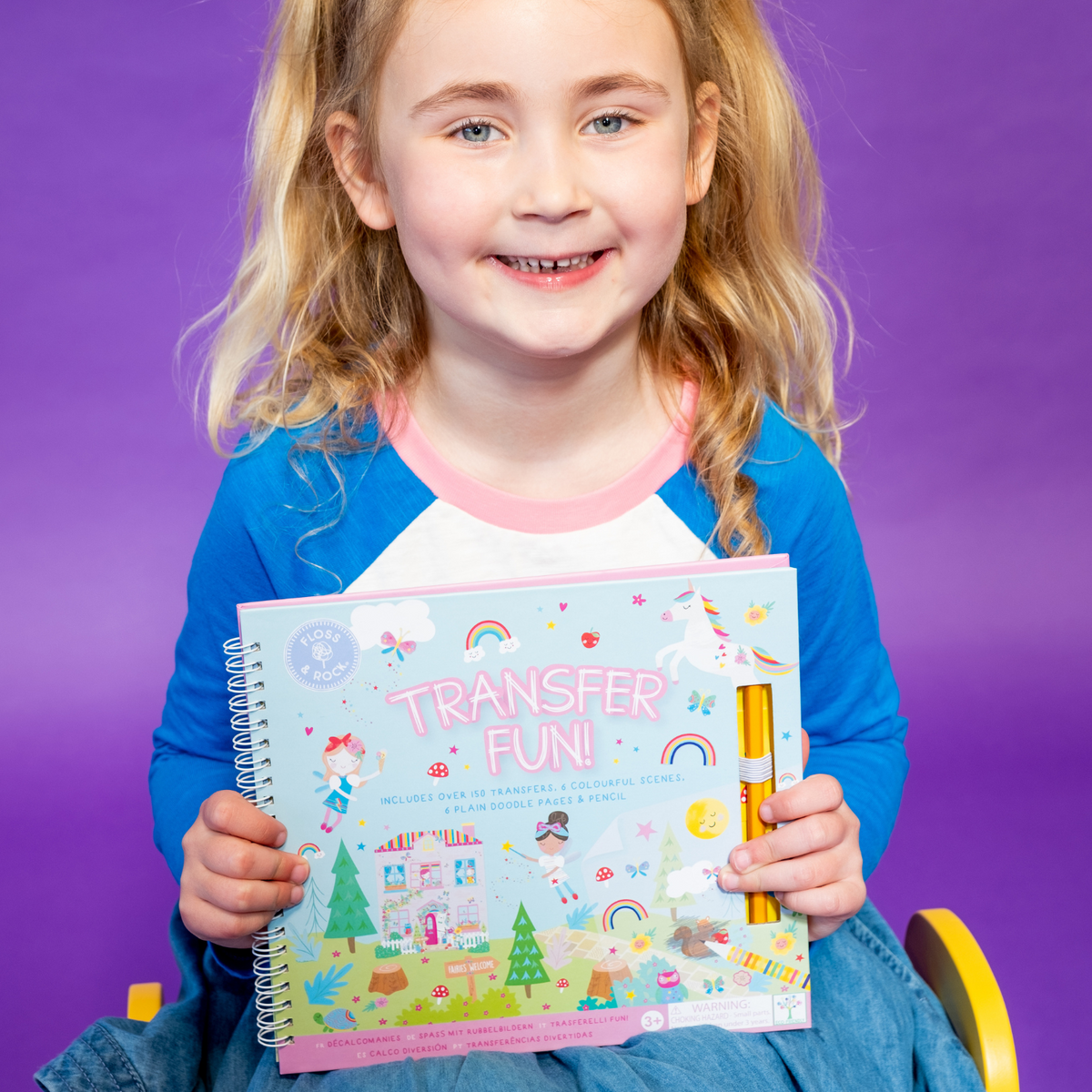 a girl holding A spiral bound book titled &#39;TRANSFER FUN&#39; featuring rainbow fairy themed transfers, with colorful illustrations on the cover including fairies and rainbows, accompanied by a yellow pencil.
