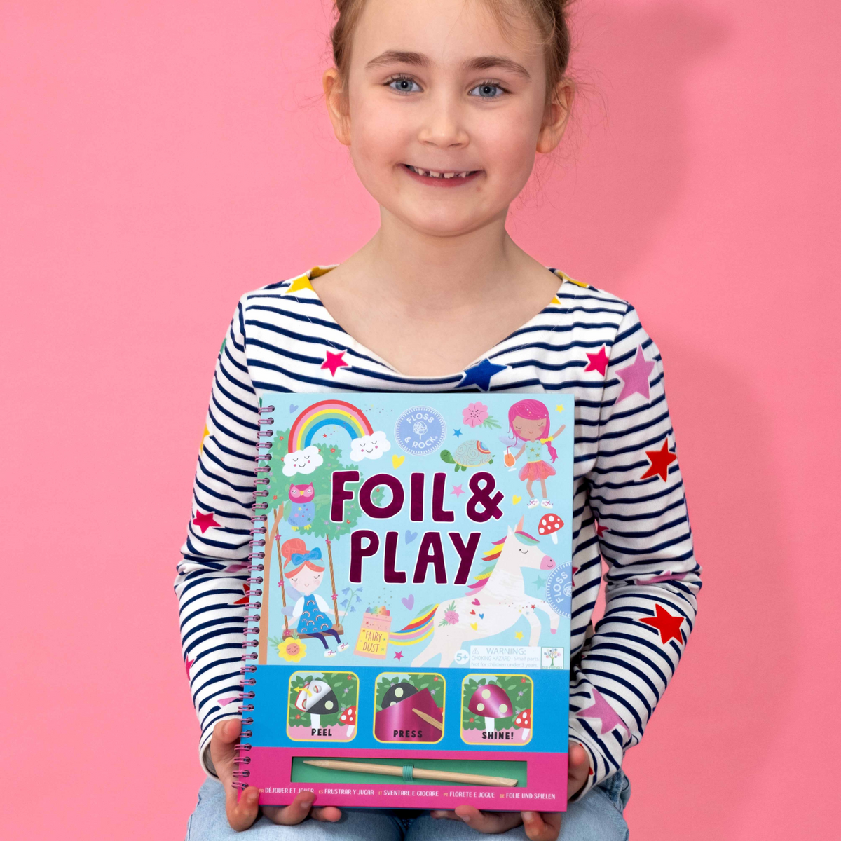 a girl holding A spiral bound book titled &#39;FOIL &amp; PLAY&#39; with illustrations of fairies and rainbow. It includes a wooden stylus and color foiling sheets.