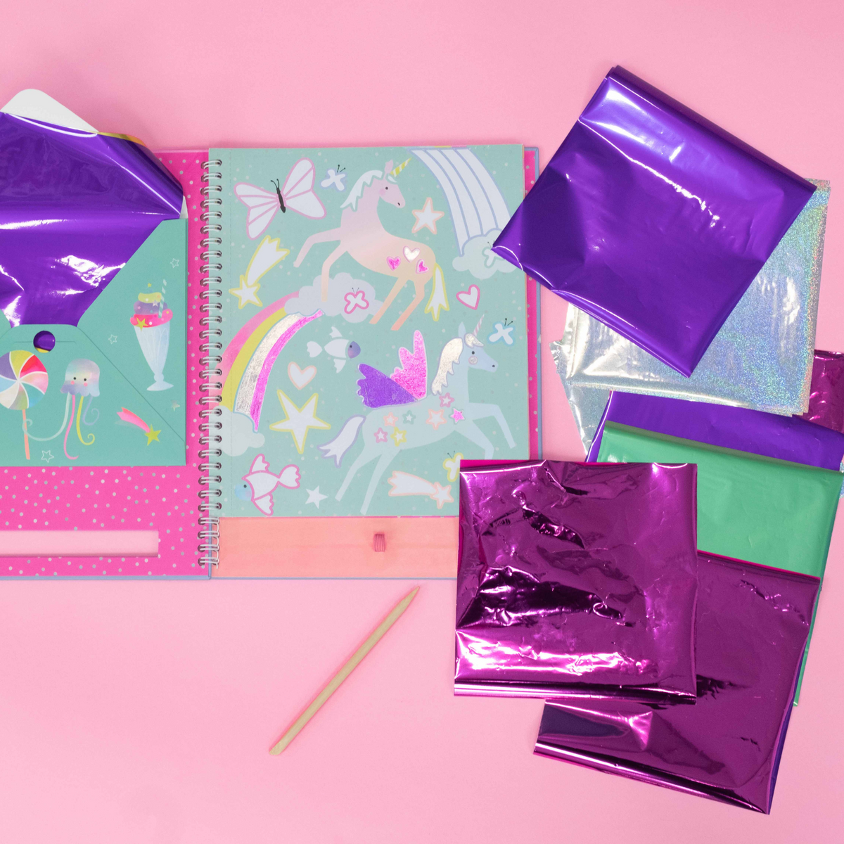 A spiral bound book titled &#39;FOIL &amp; PLAY&#39; with illustrations of fairies and rainbow. It includes a wooden stylus and color foiling sheets.