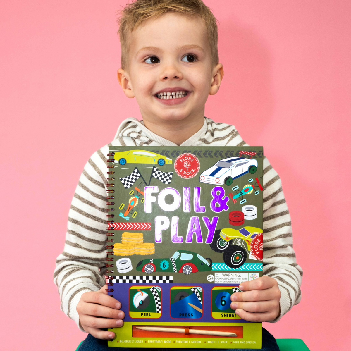 A boy holding A spiral bound book titled &#39;FOIL &amp; PLAY&#39; with colorful illustrated pages and various car designs.