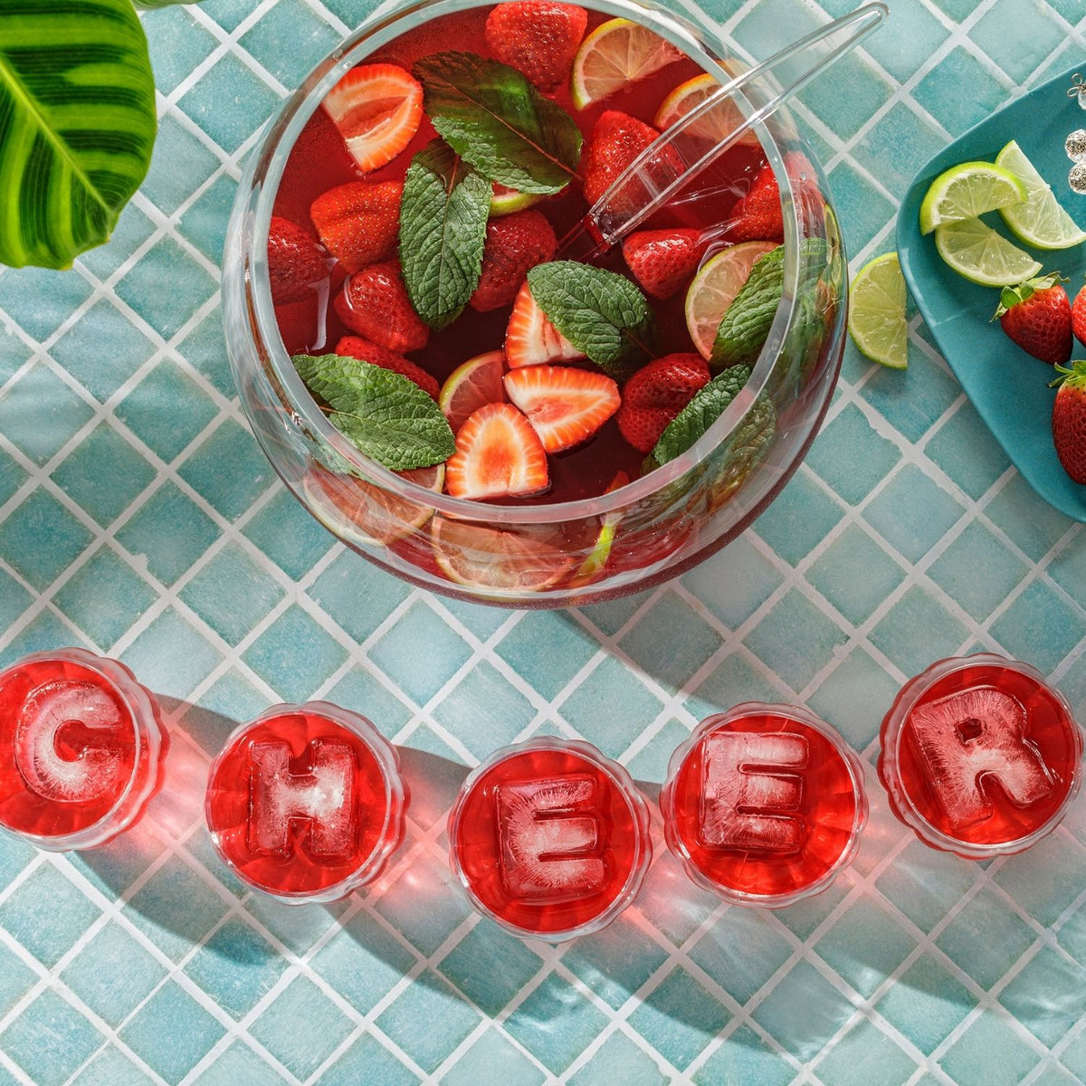 Glass punch bowl filled with strawberry lemonade, garnished with mint leaves, on a blue tiled surface with 'CHEERS' lettered ice cubes. A clear, solid ice lettersmade with ice tray mould, showing the smooth finish and sharp, modern edges.