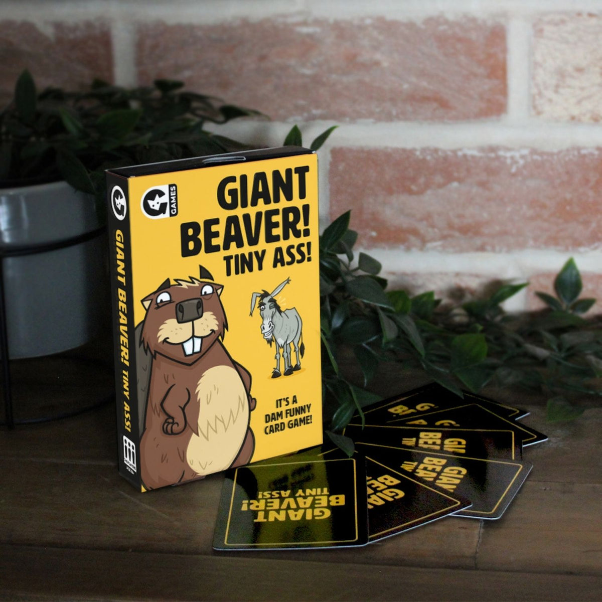 Card game packaging for 'Giant Beaver! Tiny Ass!' with cards on a wooden surface.