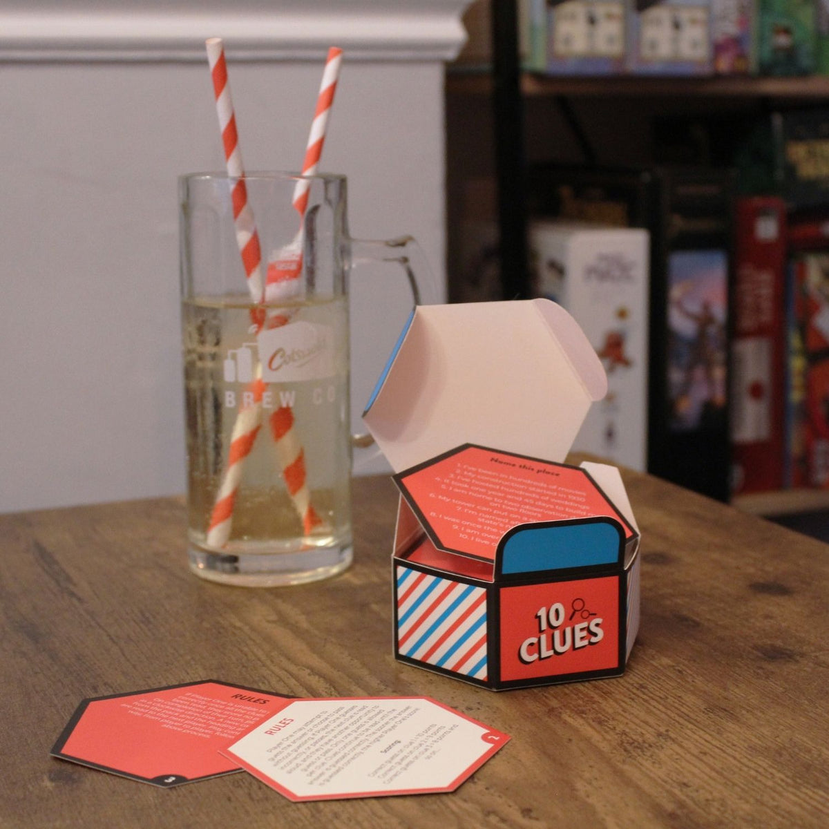 A travel-sized card game called &#39;10 Clues&#39; with a blue, red, and white geometric design on a wooden table next to drinks