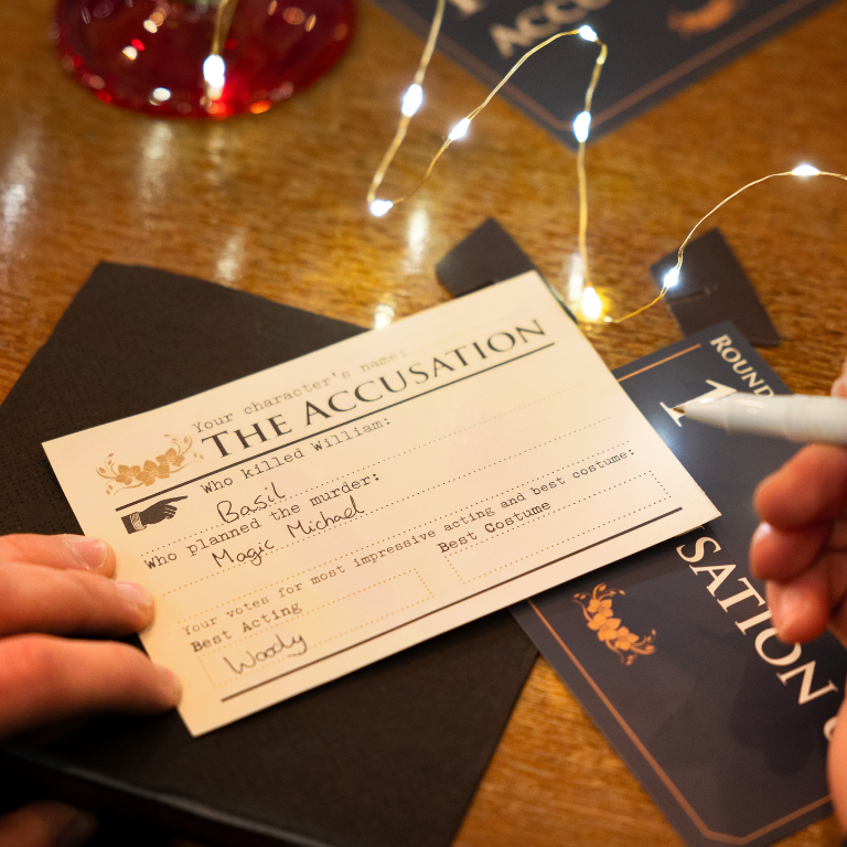Card titled 'The Accusation' held by a person with decorative lights in the background for a murder mystery game night 