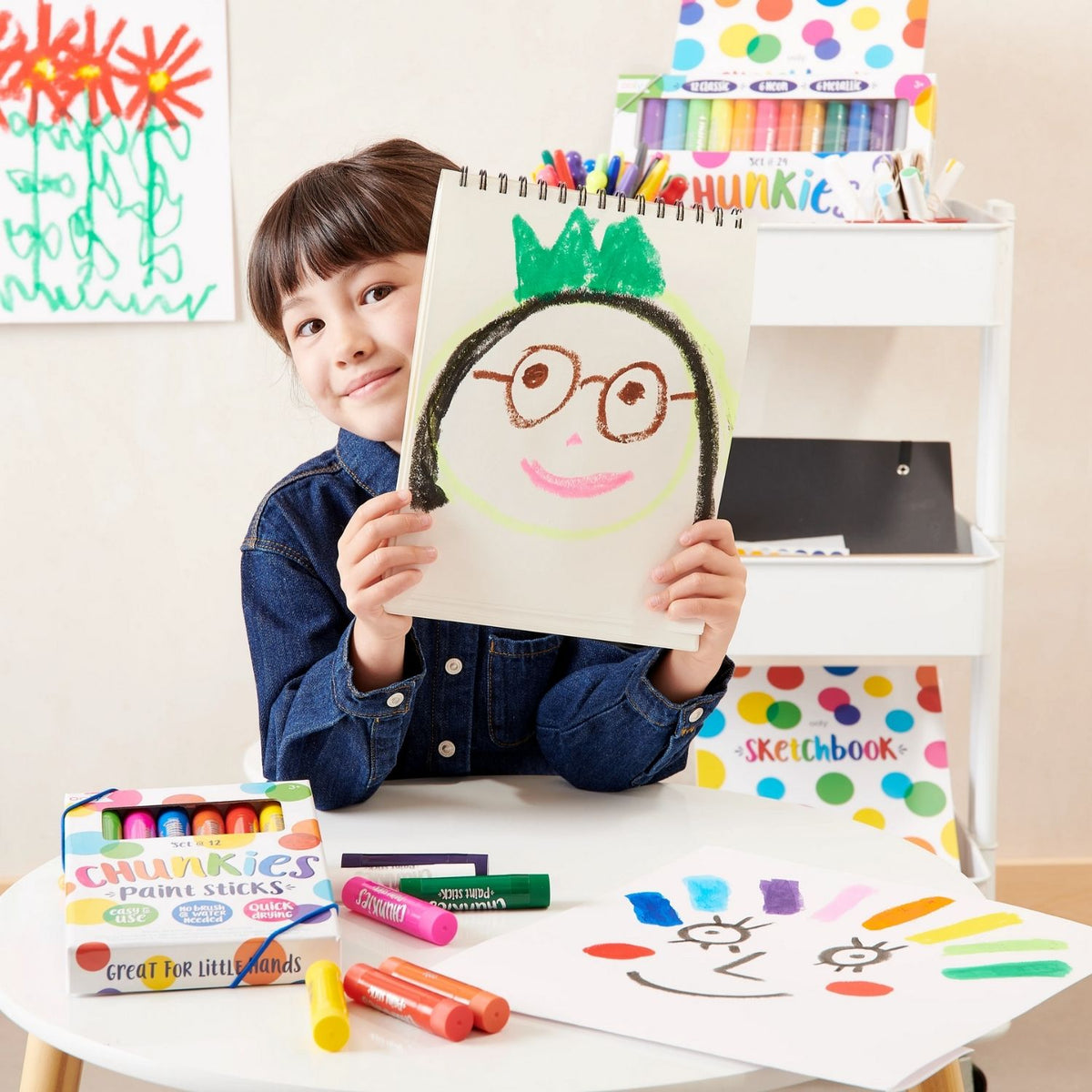 Child holding a drawing of a face with a green hat and glasses, surrounded by her chunkies paint sticks
