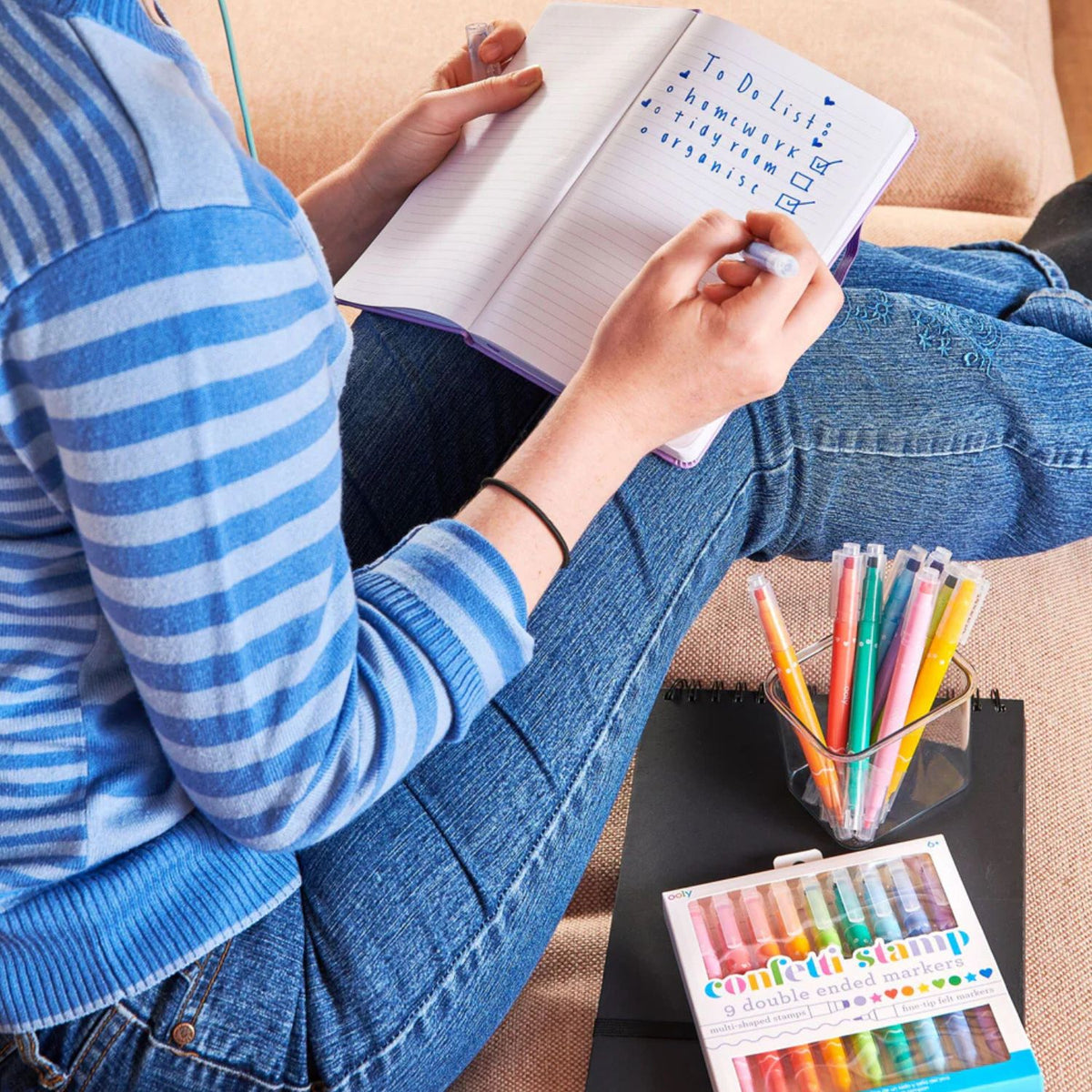 Person sitting on a couch with a notebook and using the colourful markers and stamps for journaling.