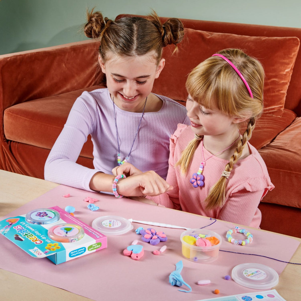 Two young girls making jewelry, smiling while wearing a completed Blissful Blooms necklace and bracelet, showcasing the pride of creating personal accessories.